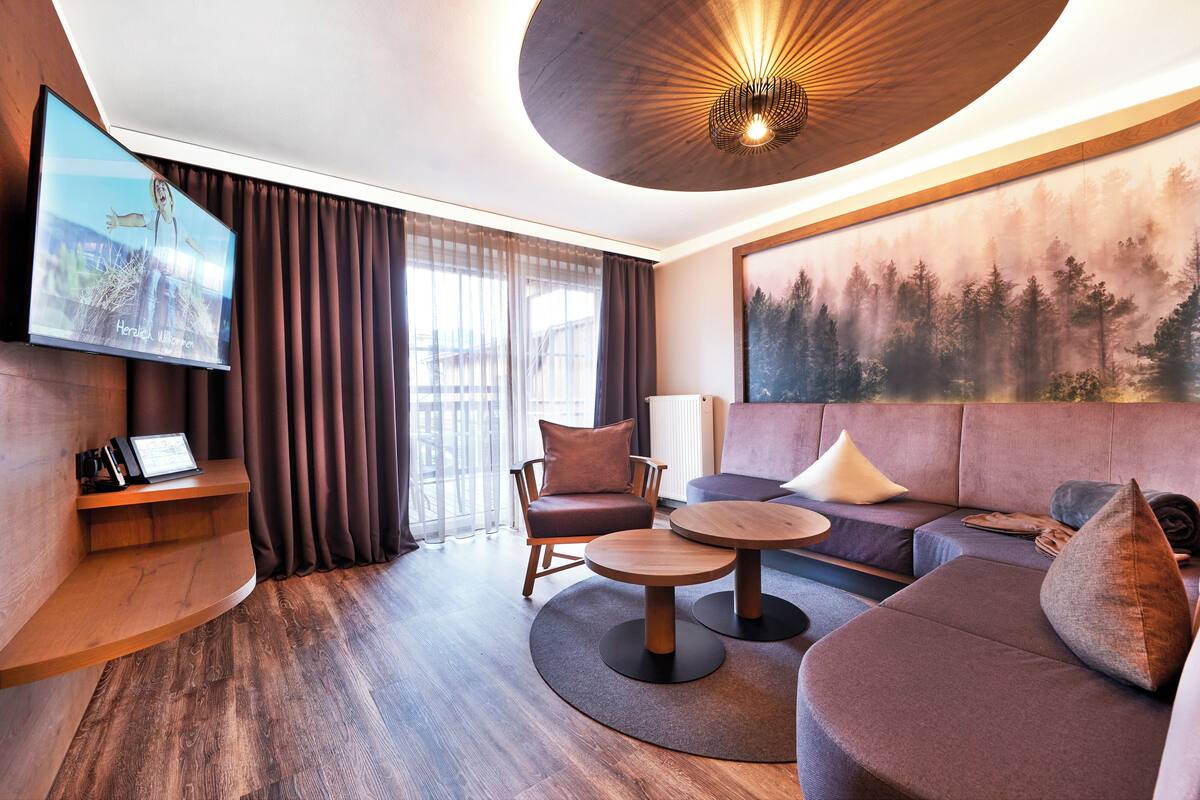 Un salon moderne comprenant un canapé, un fauteuil et une table basse, un téléviseur à écran plat et une grande fresque représentant une forêt.