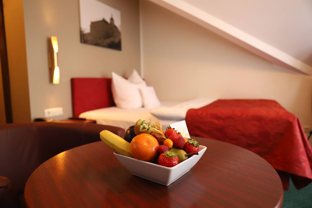 A hotel room with a bed, a red blanket, a white pillow, a fruit bowl on a table, and a wall-mounted light.