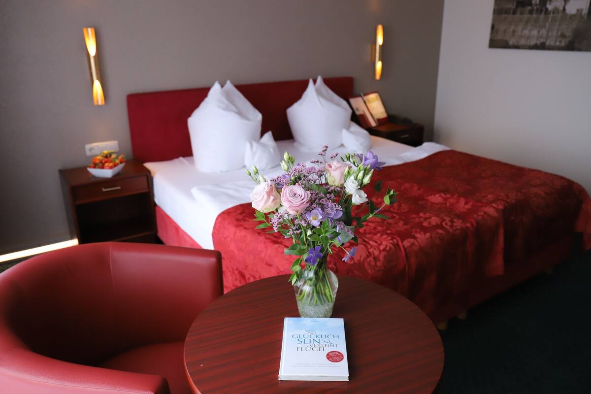 A hotel room with a neatly made bed, a red chair, a small table with a vase of flowers, and a nightstand with a bowl of fruit.