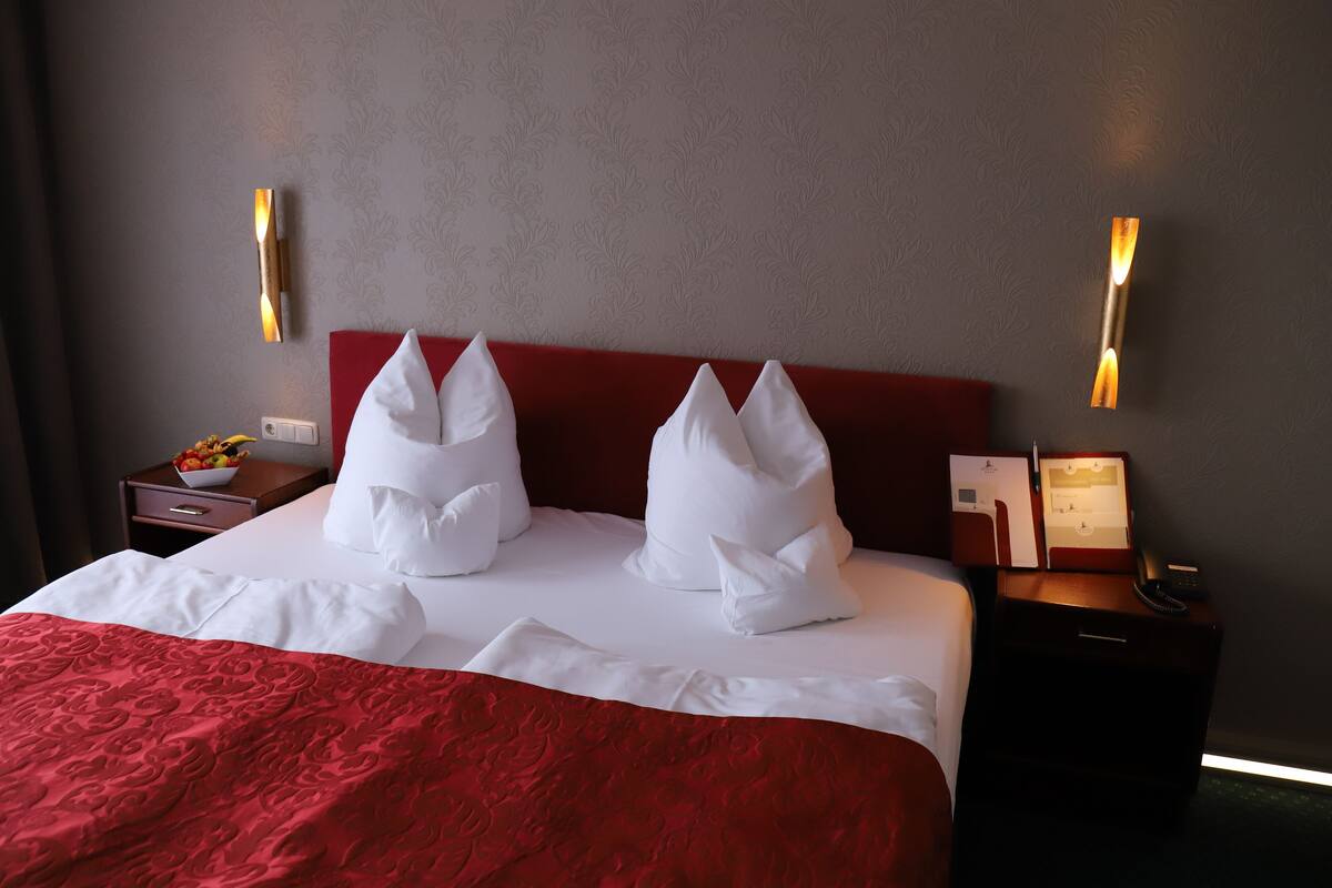 A neatly made bed with white linens and a red blanket, a wooden nightstand with a lamp, and a wall-mounted light fixture.
