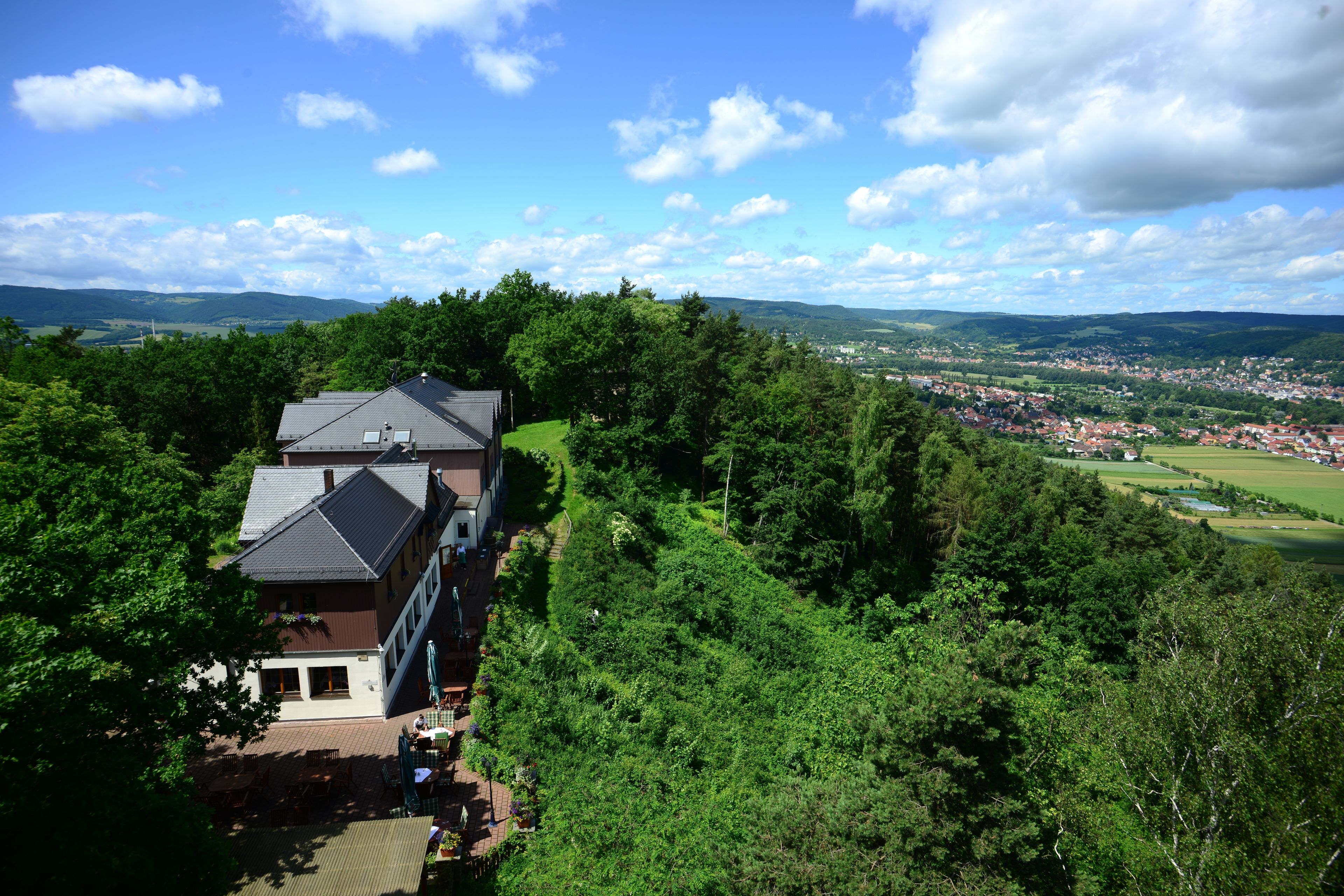 A large house with a sloped roof, surrounded by dense greenery and overlooking a valley.