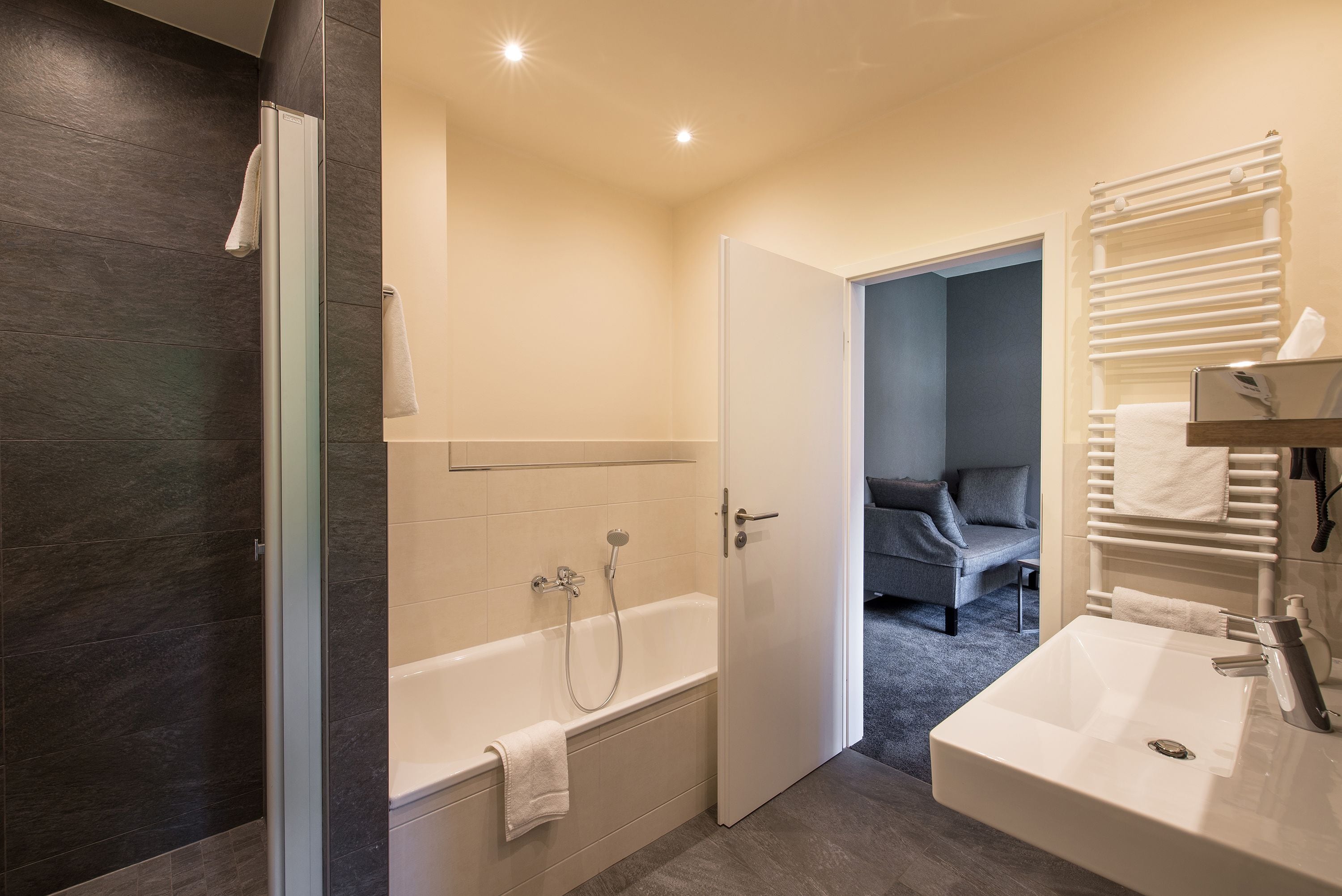 A modern bathroom with a bathtub, a shower, and a sink. There is a door leading to a bedroom with a bed and a chair.