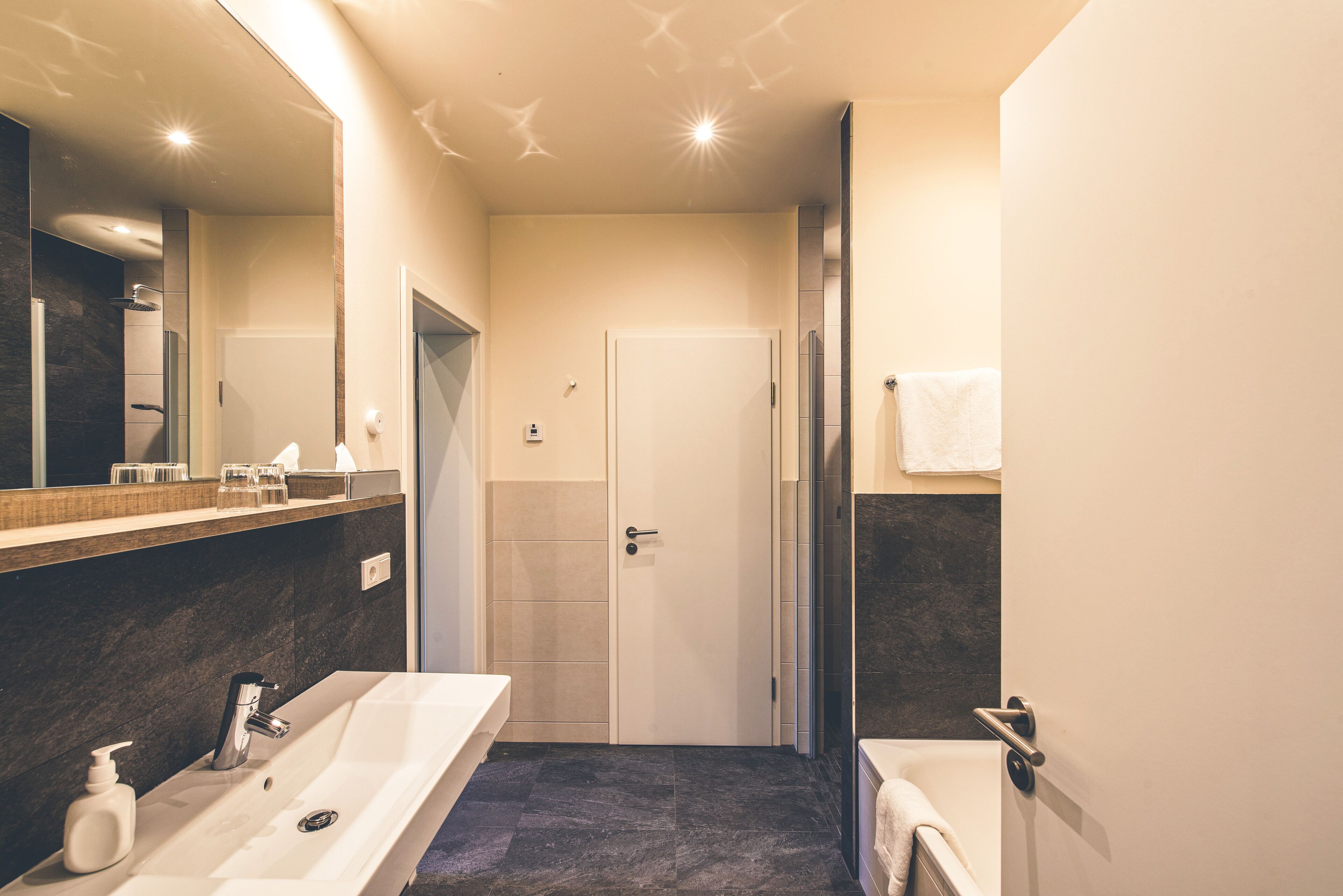 A modern bathroom with a large mirror, two sinks, a shower, and a door leading to another room.