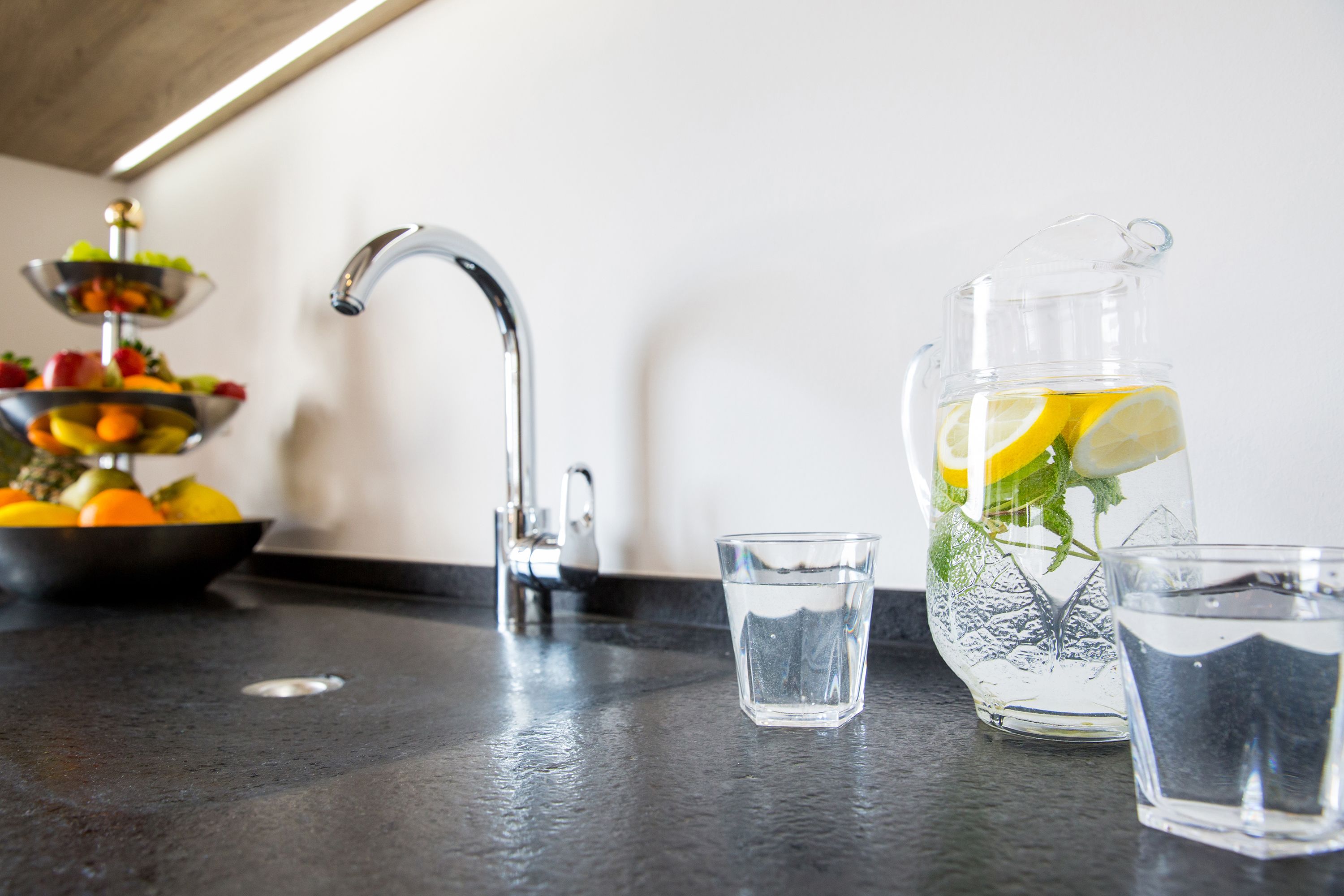 Una encimera de cocina con un frutero, una jarra de agua con rodajas de limón y dos vasos.