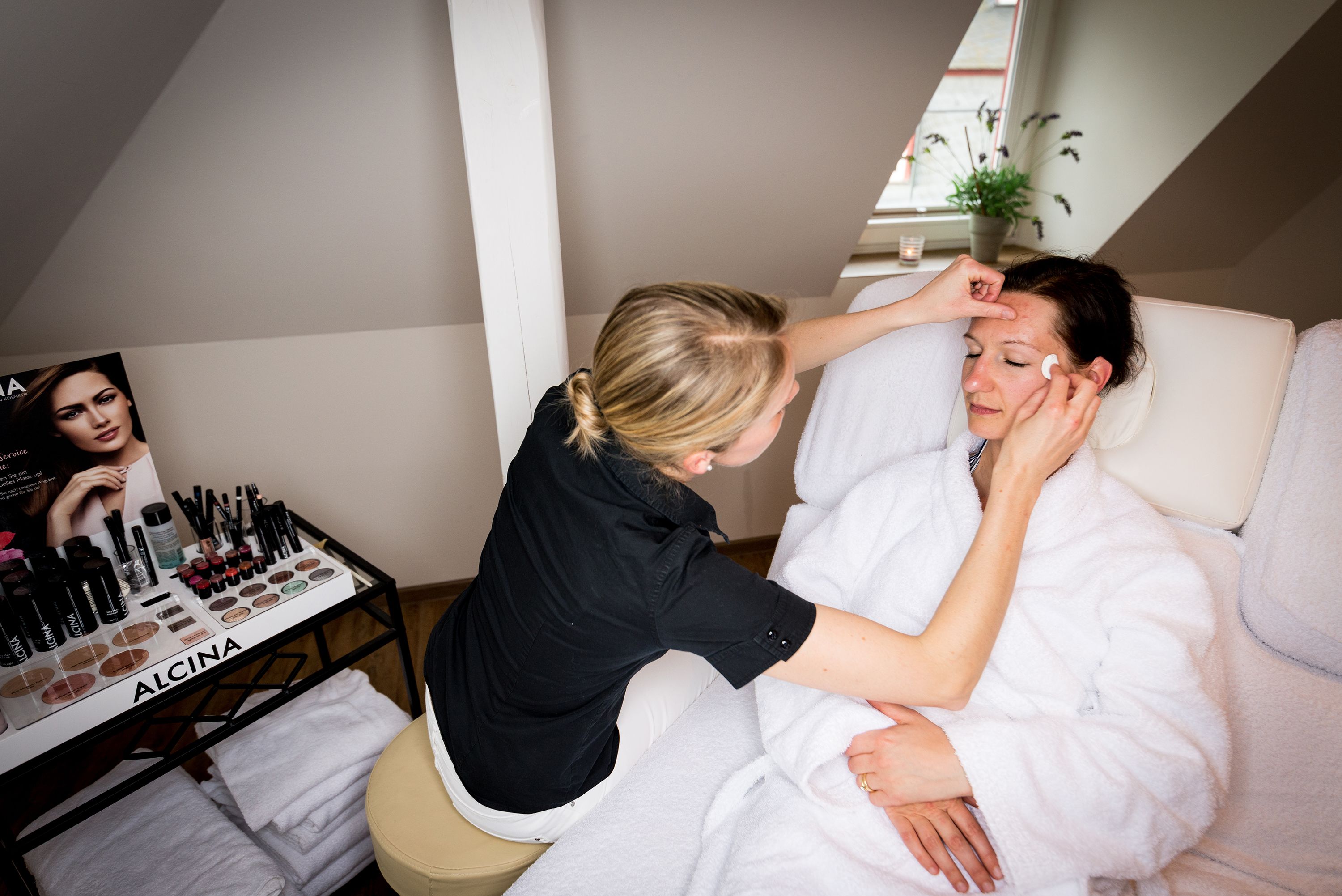 A person receiving a facial treatment in a room with a window, a table with skincare products, and a poster on the wall.