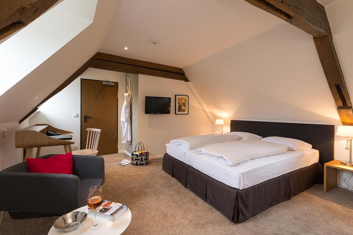 A modern hotel room with a large bed, a sofa, a small table with a glass of wine, and a TV on the wall.