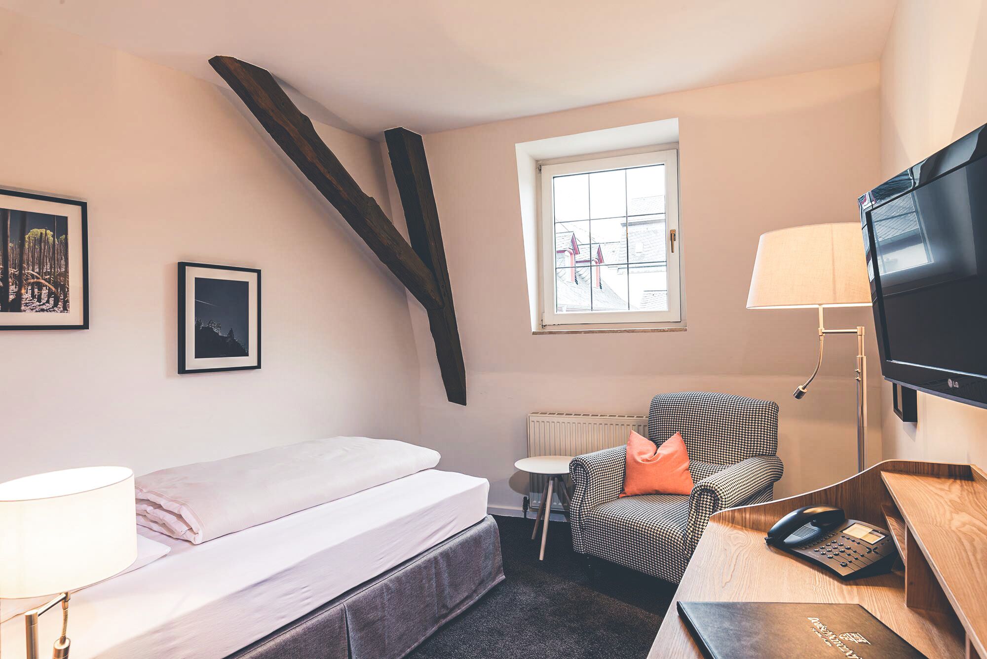 A compact hotel room with a bed, a chair, a desk with a telephone, a TV, and a window with a view of buildings.