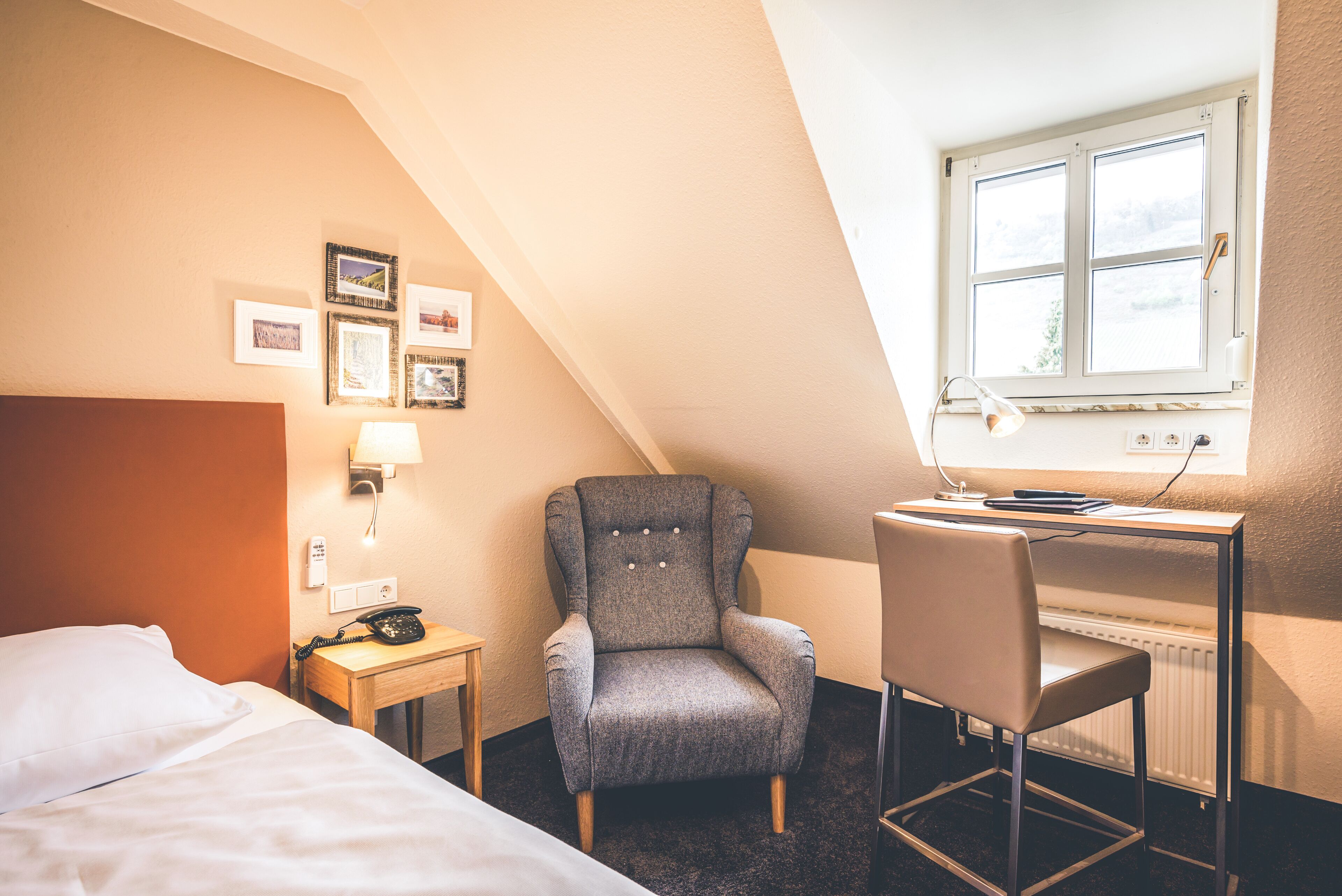 A hotel room with a bed, a desk with a chair, a telephone, and framed pictures on the wall.