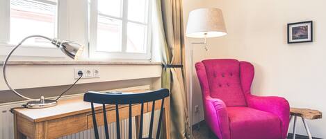 Una habitación con un escritorio de madera, una silla negra, un sillón rosa, una lámpara de pie y una ventana con cortinas.