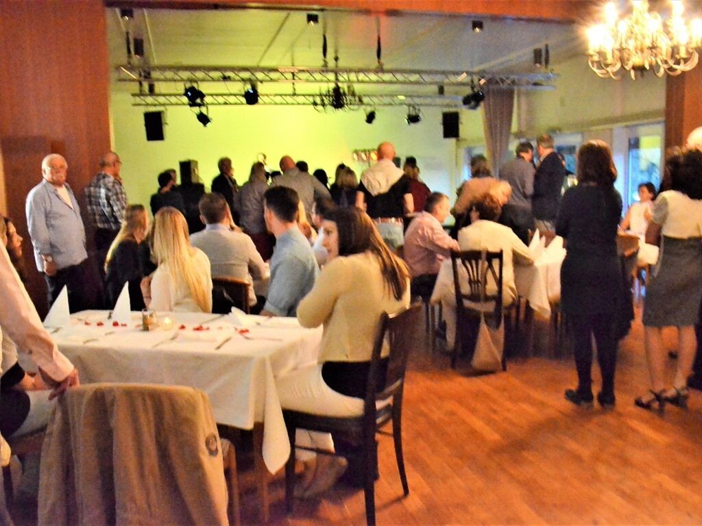 A crowded indoor event with tables set for dining, a stage with lighting equipment, and a chandelier.