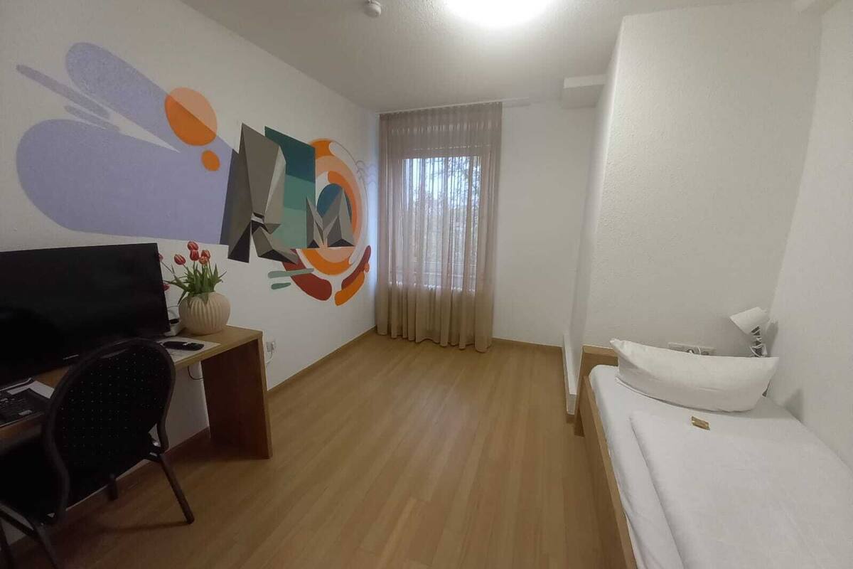 A hotel room with a bed, a desk with a computer, a chair, a TV, a wall with abstract art, and a window with curtains.