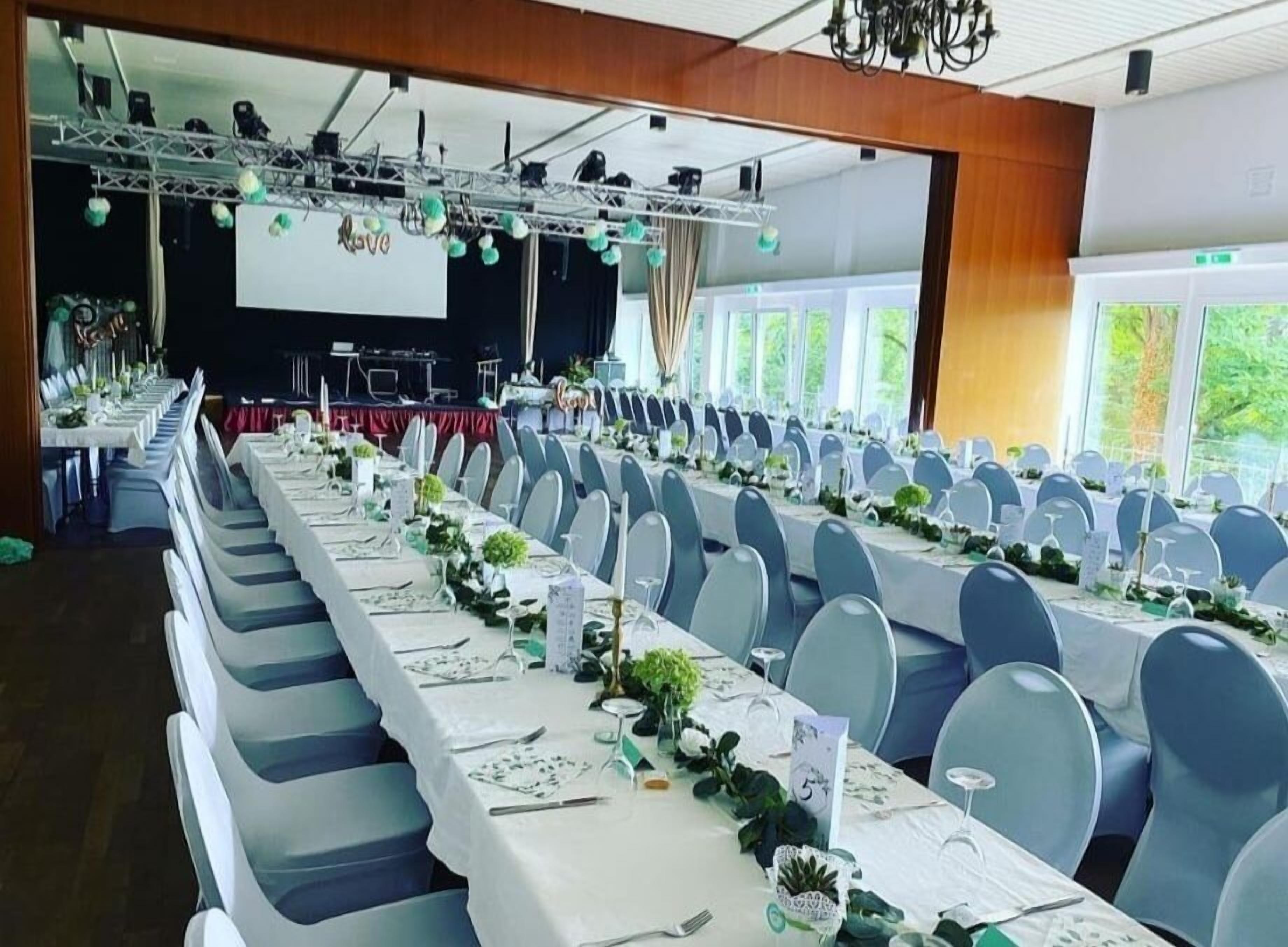 Un salón de banquetes con largas mesas dispuestas para un evento formal, equipado con sillas, mantelería y un escenario con iluminación.