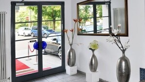 En hotellobby med en röd matta, tre metallvaser med blommor, ett stort fönster med utsikt över parkerade bilar och ett element på väggen.