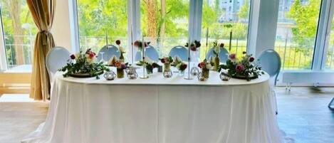 Una mesa redonda con mantel blanco, decorada con centros de mesa florales y candelabros, con un fondo de grandes ventanales que dan a vistas de zonas verdes y edificios.