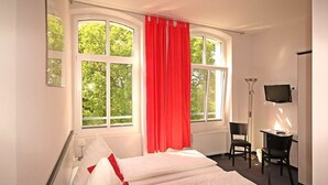 Habitación de hotel con cama, escritorio, silla, televisor y ventanas grandes con cortinas.
