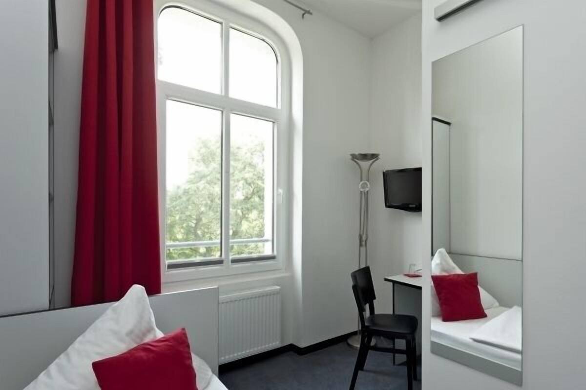 Ett hotellrum med en säng, ett skrivbord, en stol, en TV och ett fönster med utsikt över träd.