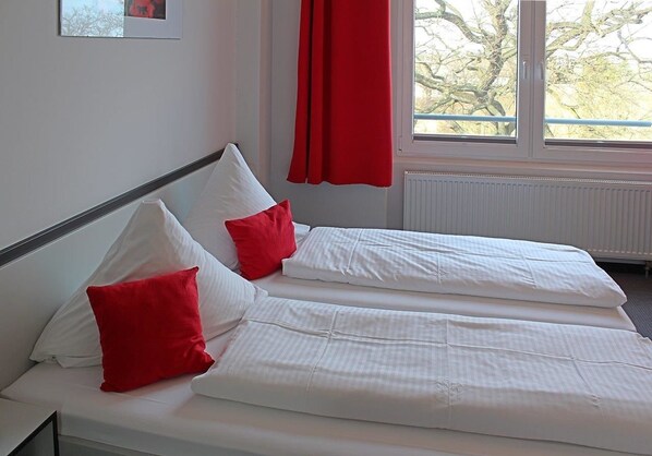 A double bed with white bedding and red pillows, a window with a red curtain, and a radiator below.