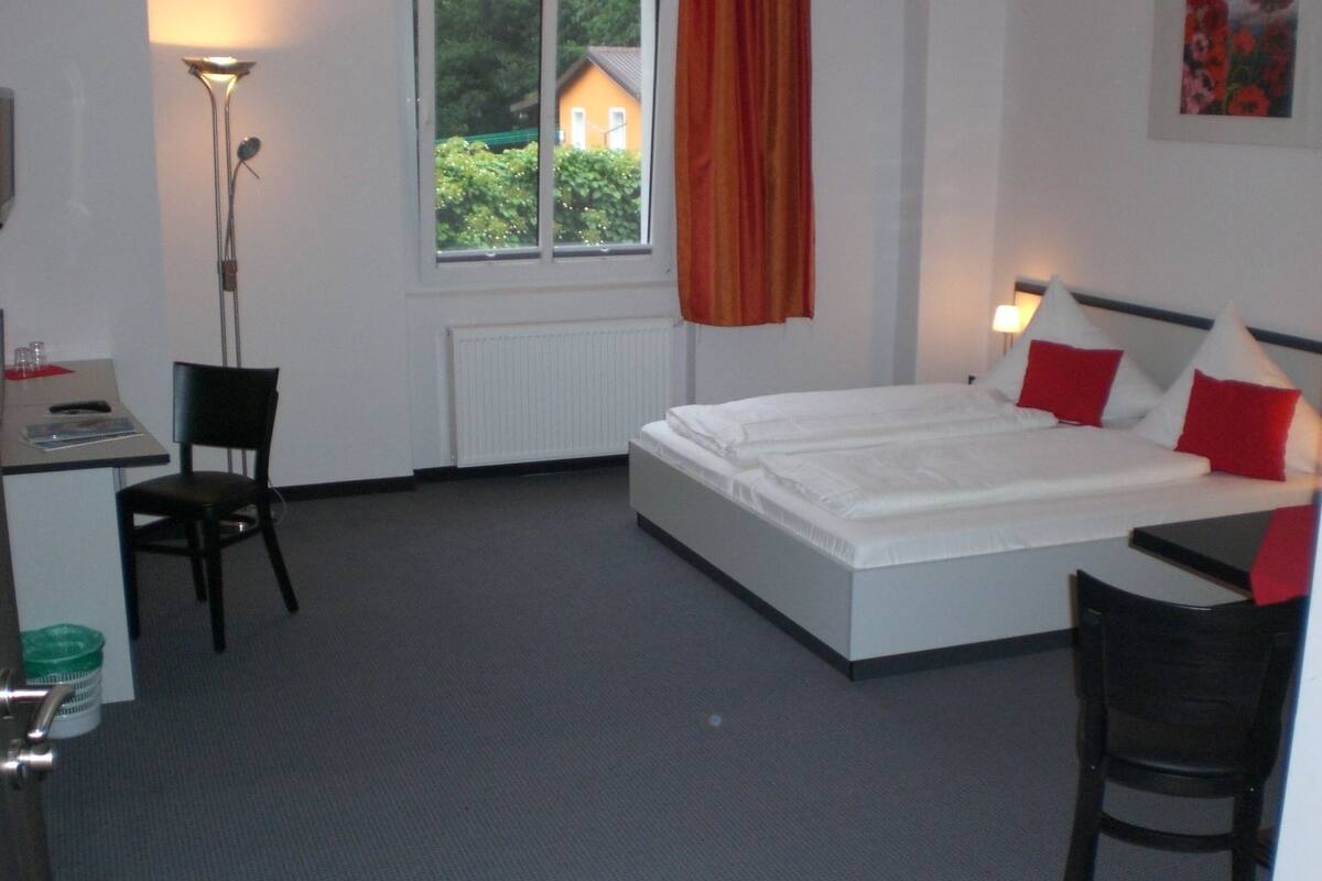 Ett hotellrum med en säng, ett skrivbord, en stol, ett element, ett fönster med utsikt över ett hus och en lampa.