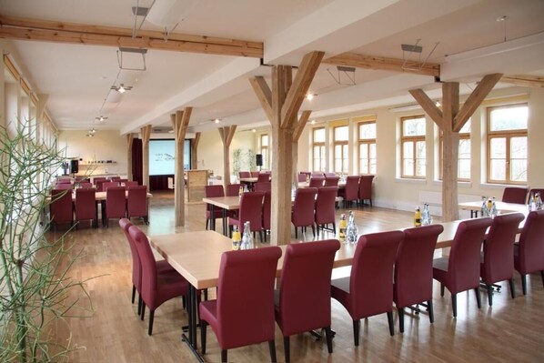 Un vaste espace dédié à la salle à manger, avec des poutres en bois, de grandes fenêtres et des chaises recouvertes d’un tissu rouge.