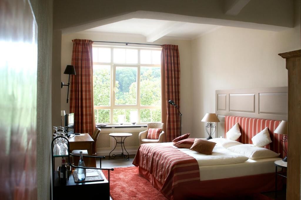 A hotel room with a large bed, a small table, a chair, and a window with curtains.
