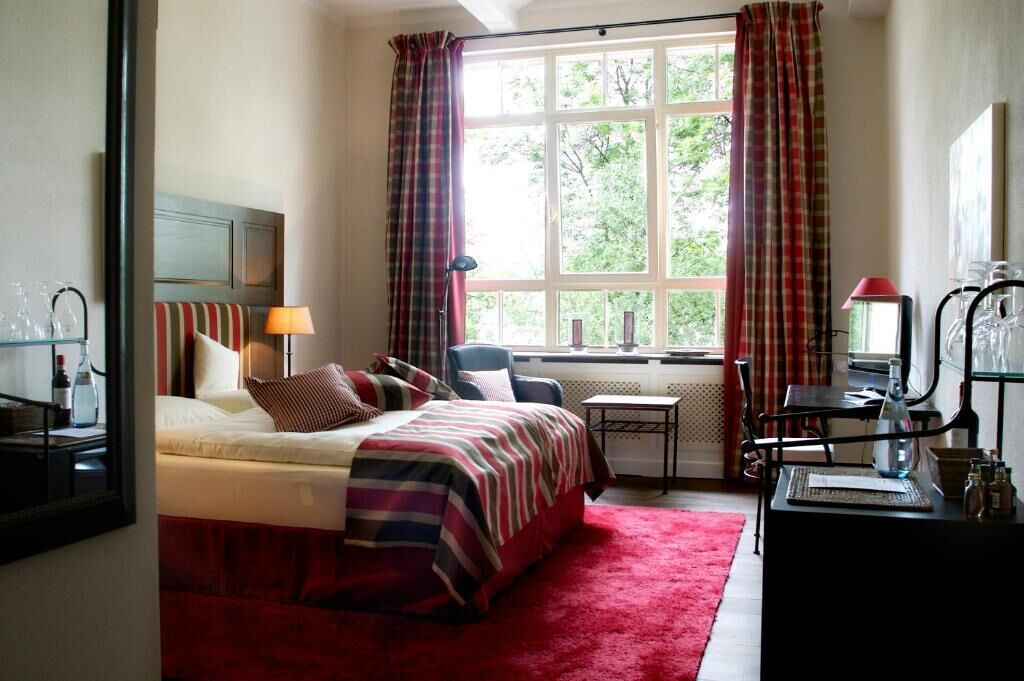 Habitación de hotel con cama, escritorio, silla, ventana con cortinas y vista a zonas verdes.