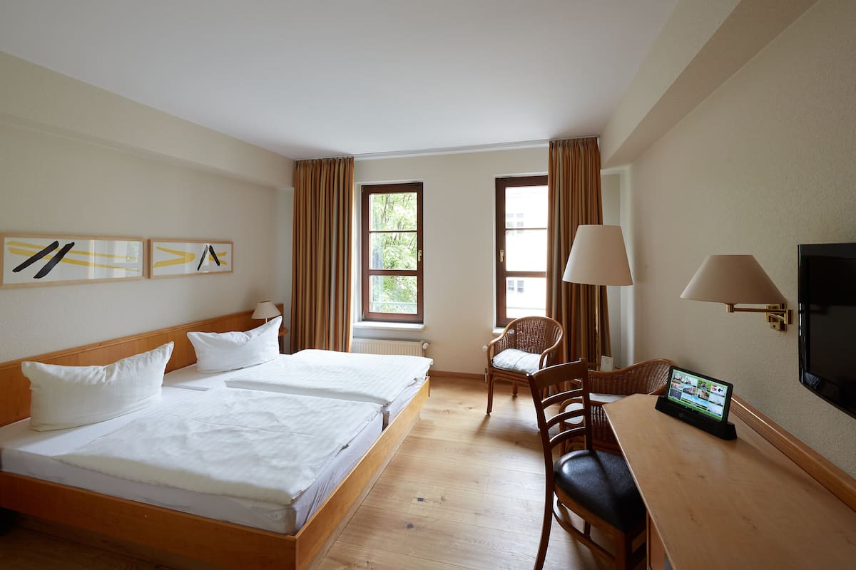 A hotel room with a double bed, a desk with a TV, and two windows with curtains.