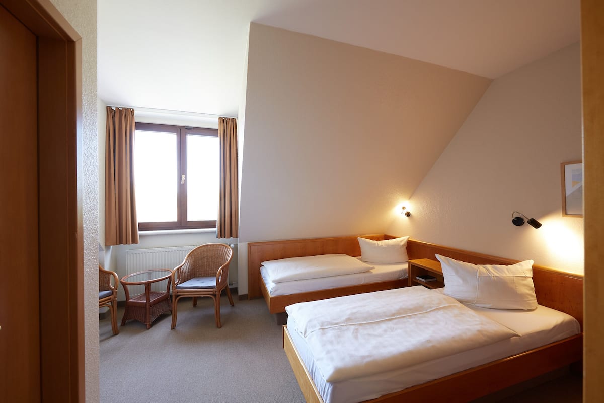 A hotel room with two single beds, a chair, a small table, and a window with curtains.