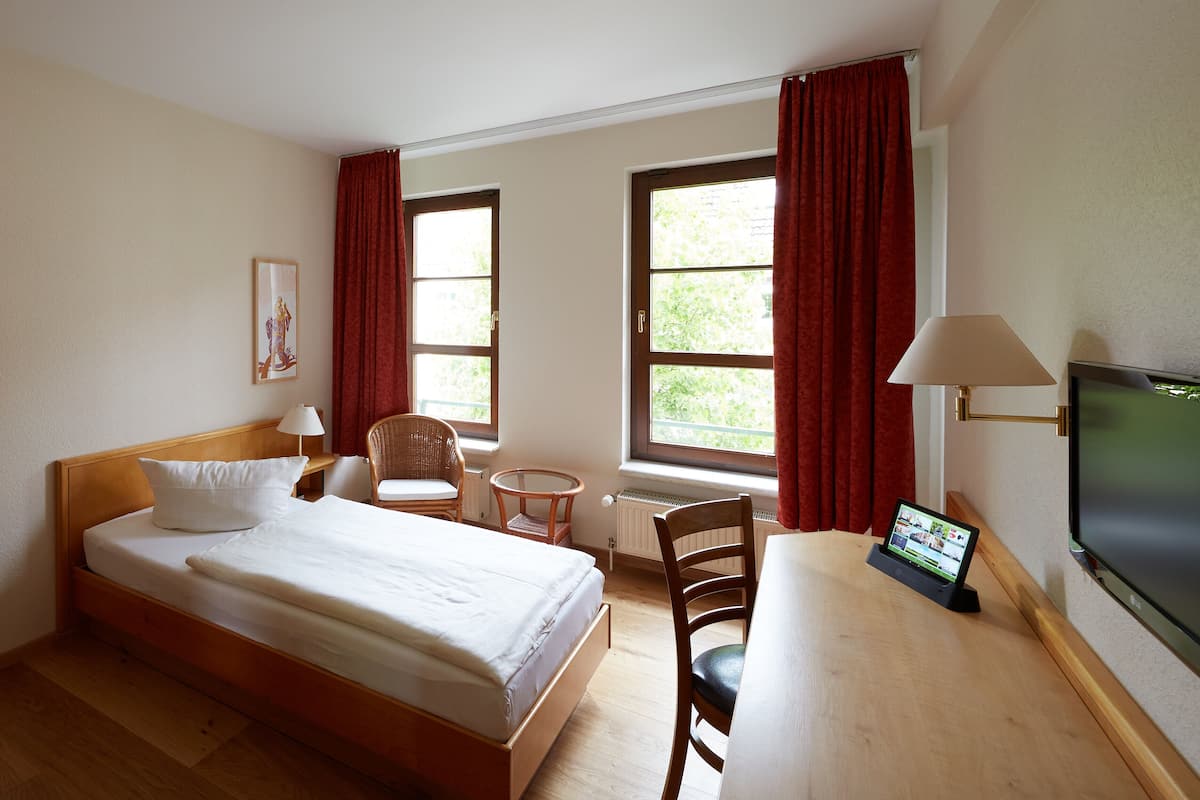 A hotel room with a bed, a desk, a chair, a TV, and two windows with curtains.