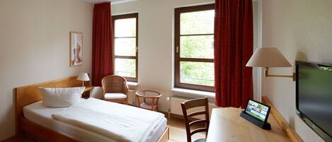 A hotel room with a bed, a desk, a chair, a TV, and two windows with curtains.
