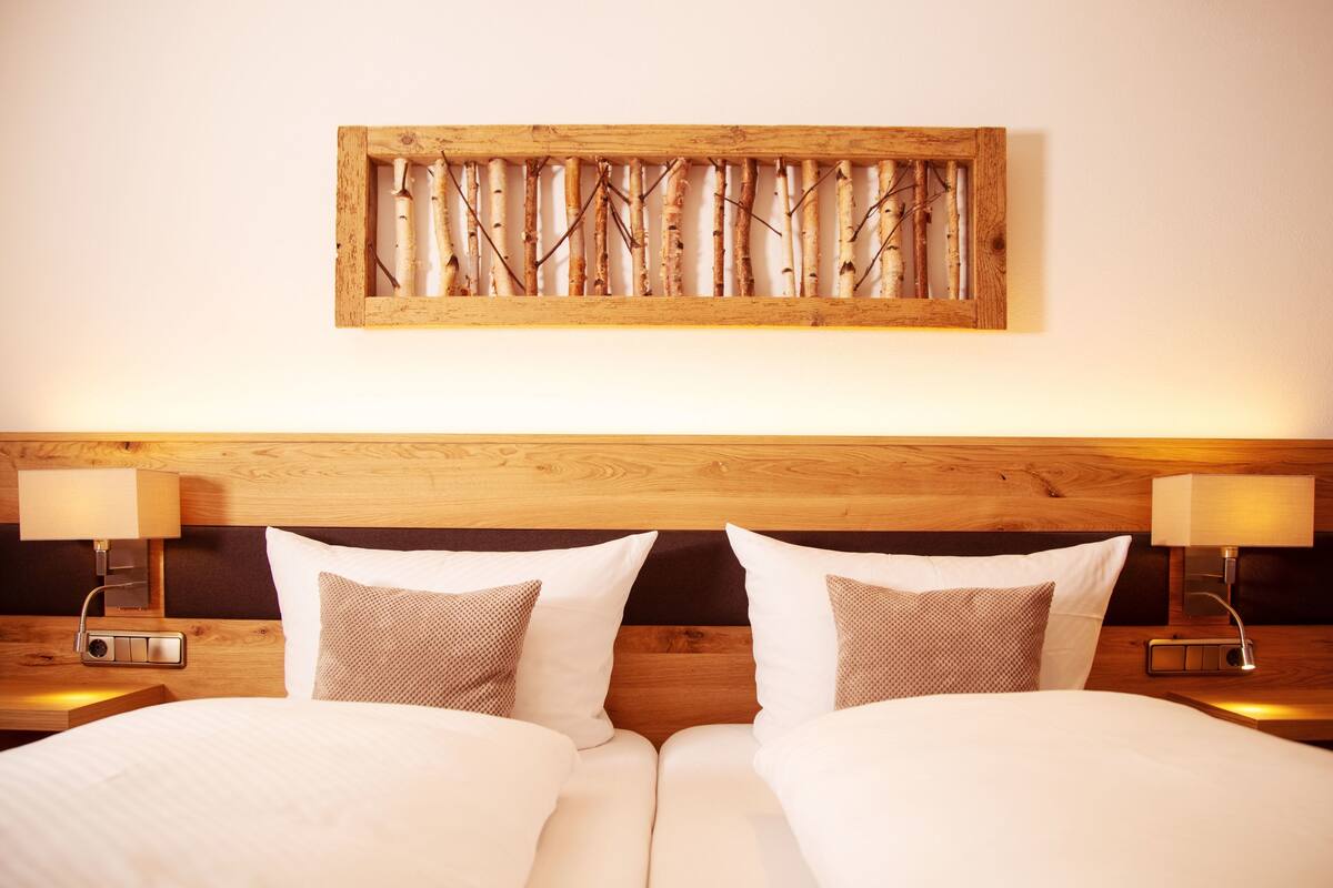 A hotel room with a wooden headboard, two beds with white bedding, two bedside lamps, and a wall-mounted artwork of branches.