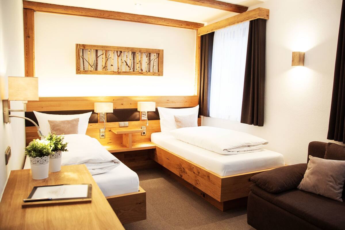 A hotel room with a wooden headboard, two beds, a wooden nightstand, a bedside table, a small potted plant, a sofa, and a wall-mounted light fixture.