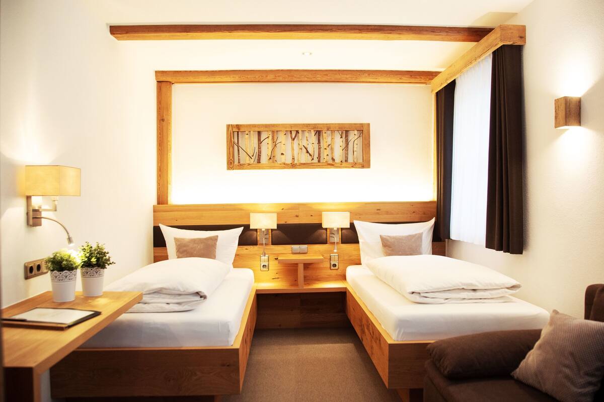 A hotel room with two single beds, a wooden headboard, bedside lamps, a small table with a lamp, a potted plant, and a sofa.