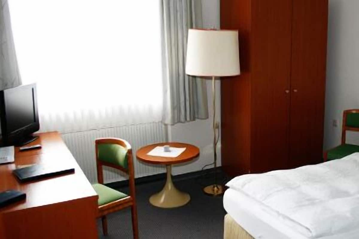 A hotel room with a bed, a desk with a computer, a chair, a small round table, and a lamp.