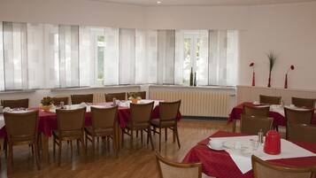 Un comedor con varias mesas y sillas, ventanas amplias con cortinas translúcidas y un radiador en la pared.