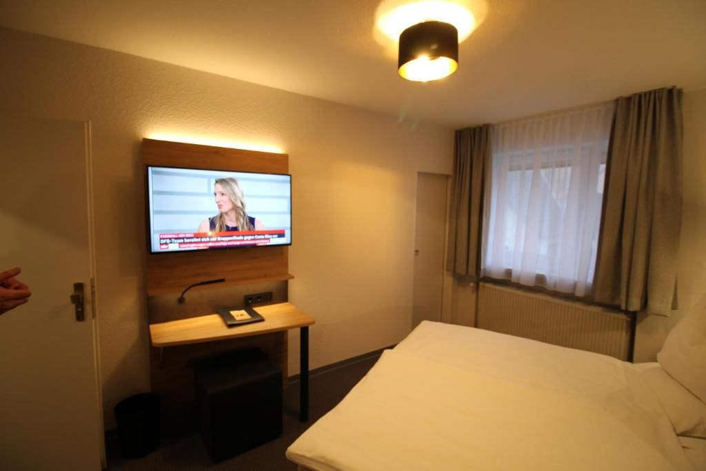 A hotel room with a bed, a desk with a chair, a television mounted on the wall, and a window with curtains.