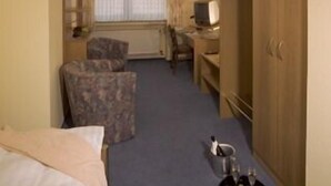 Une chambre d’hôtel de taille réduite, comprenant un lit, un petit bureau, une chaise, une armoire et une fenêtre avec des rideaux.