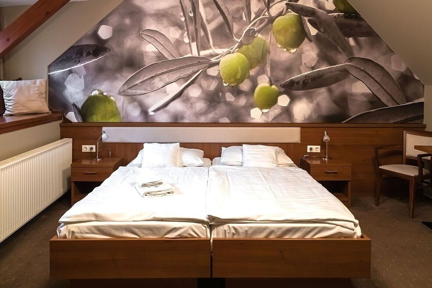 A double bed with white linens, a wooden headboard, two bedside tables with lamps, and a wall mural of leaves and fruits.