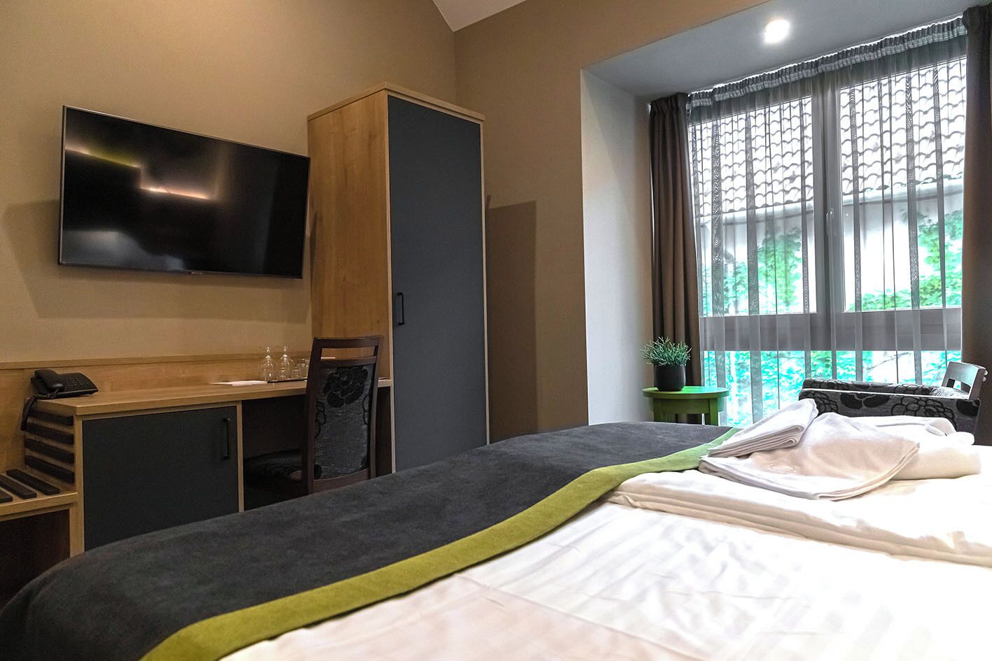 A hotel room with a bed, a TV mounted on the wall, a desk with a chair, and a window with curtains.