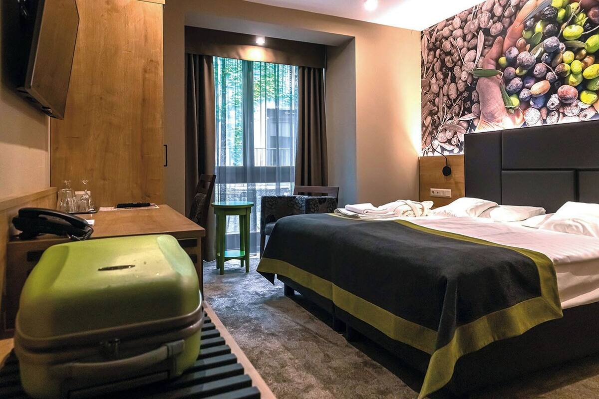 A hotel room with a bed, a desk, a chair, a green suitcase, and a window with curtains.