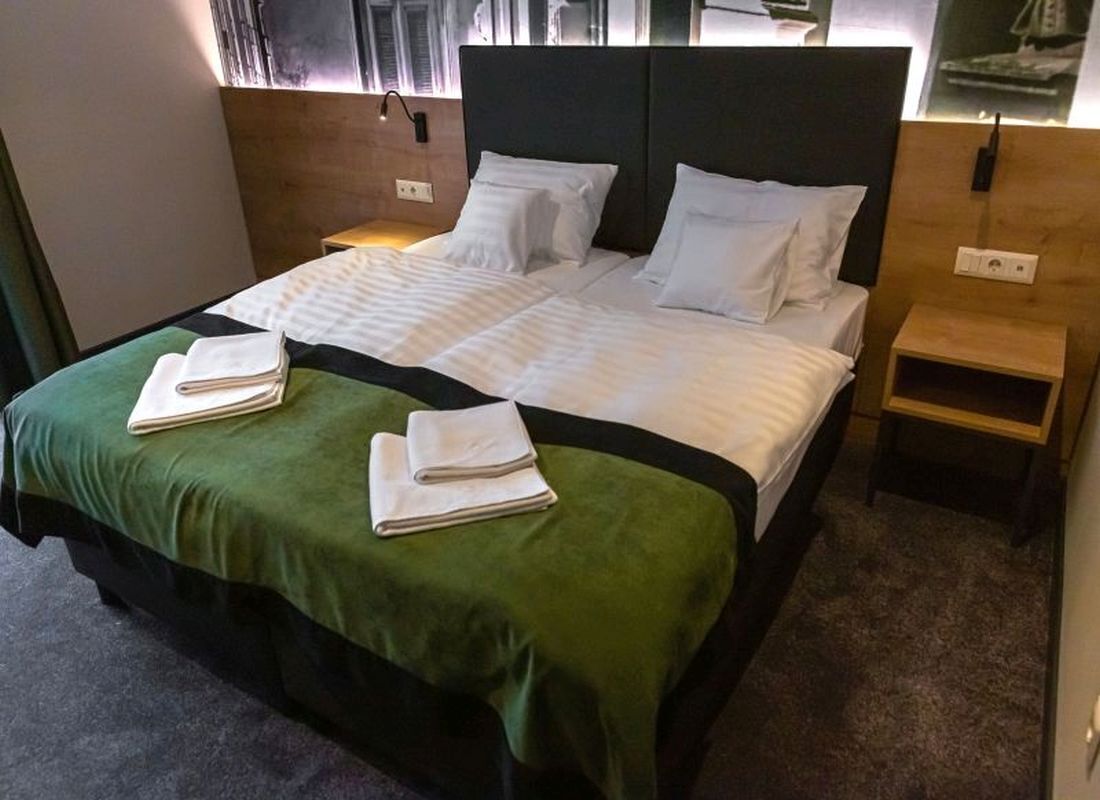 Una cama bien hecha con una colcha verde y dos toallas dobladas, un cabecero de madera y una mesita de noche de madera con una lámpara.