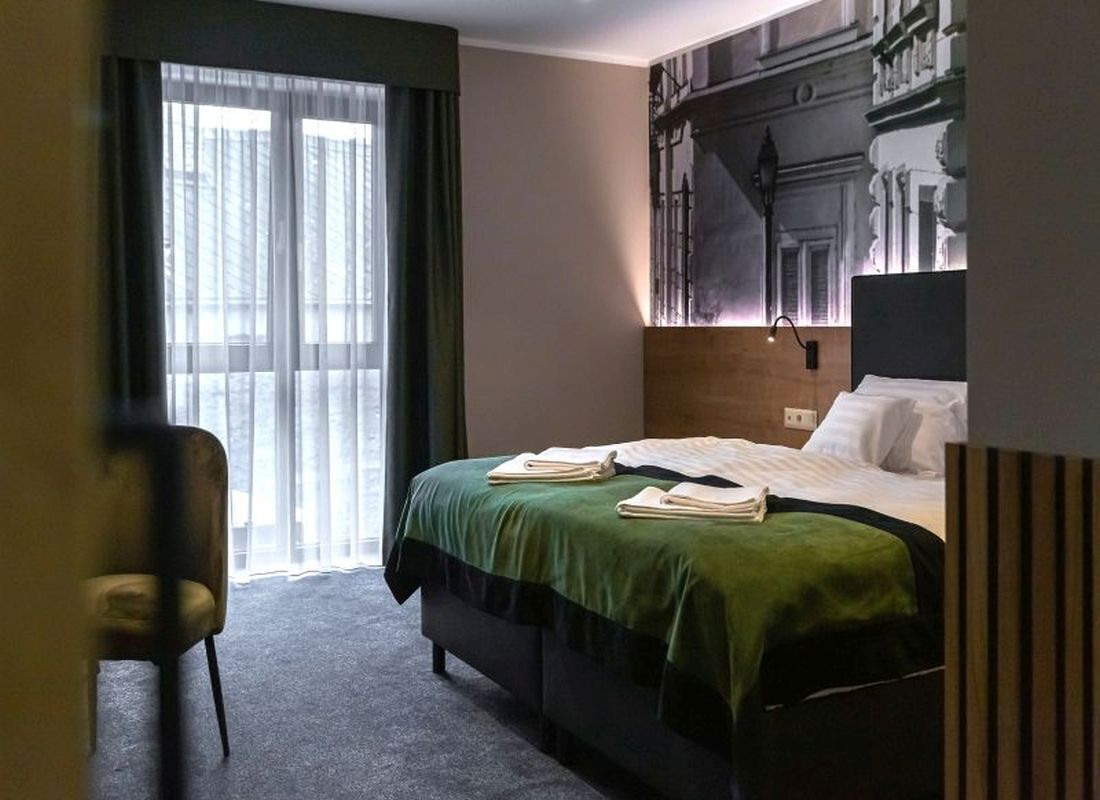 A hotel room with a bed, a chair, a large window with curtains, and a wall with architectural details.