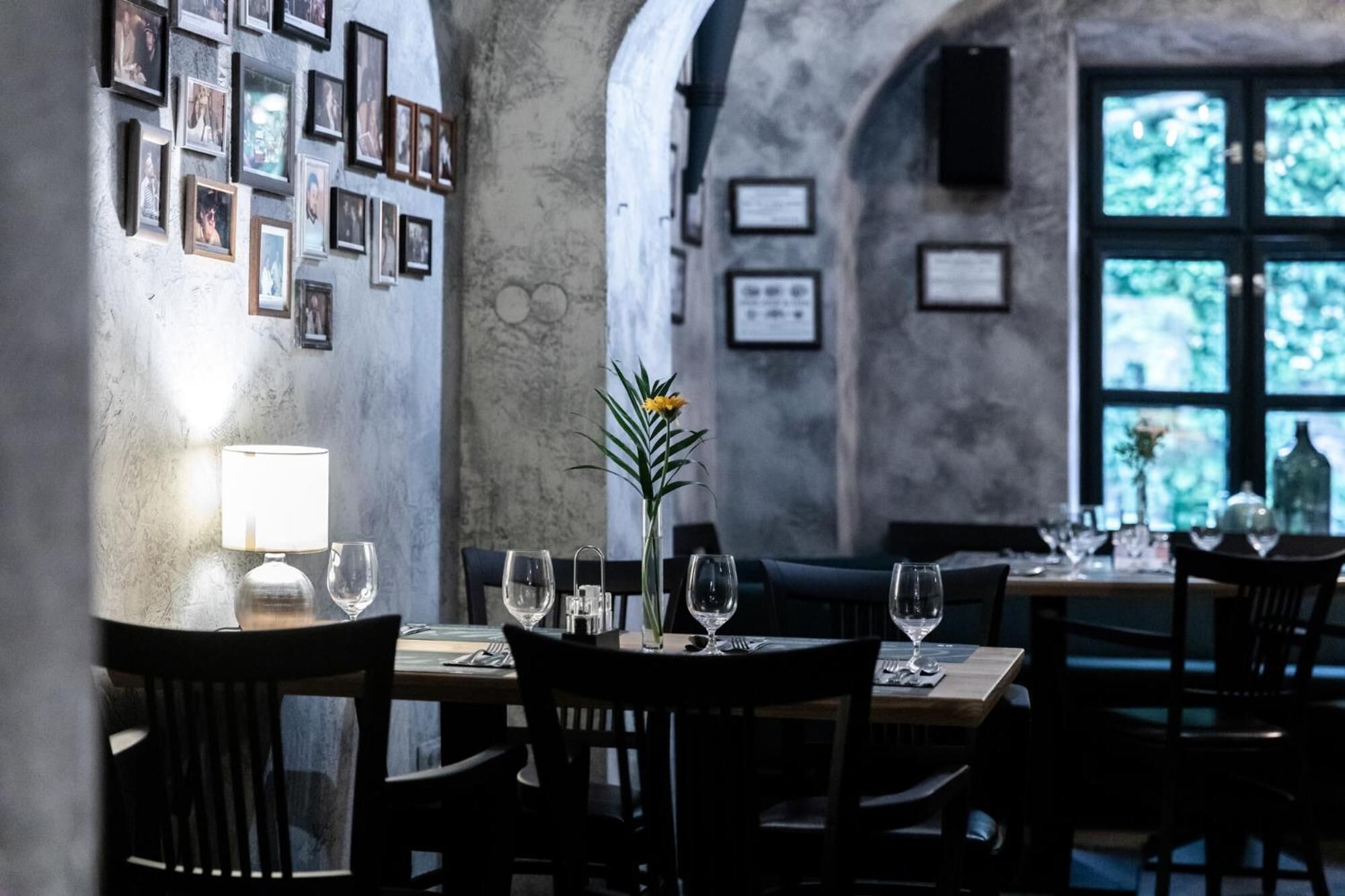 A dimly lit restaurant with tables set for dining, framed pictures on the walls, and a window with a view of greenery.