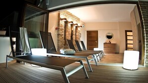 A modern room with a wooden floor, a brick wall, and a large window. There are several lounge chairs and a small table with a lamp.