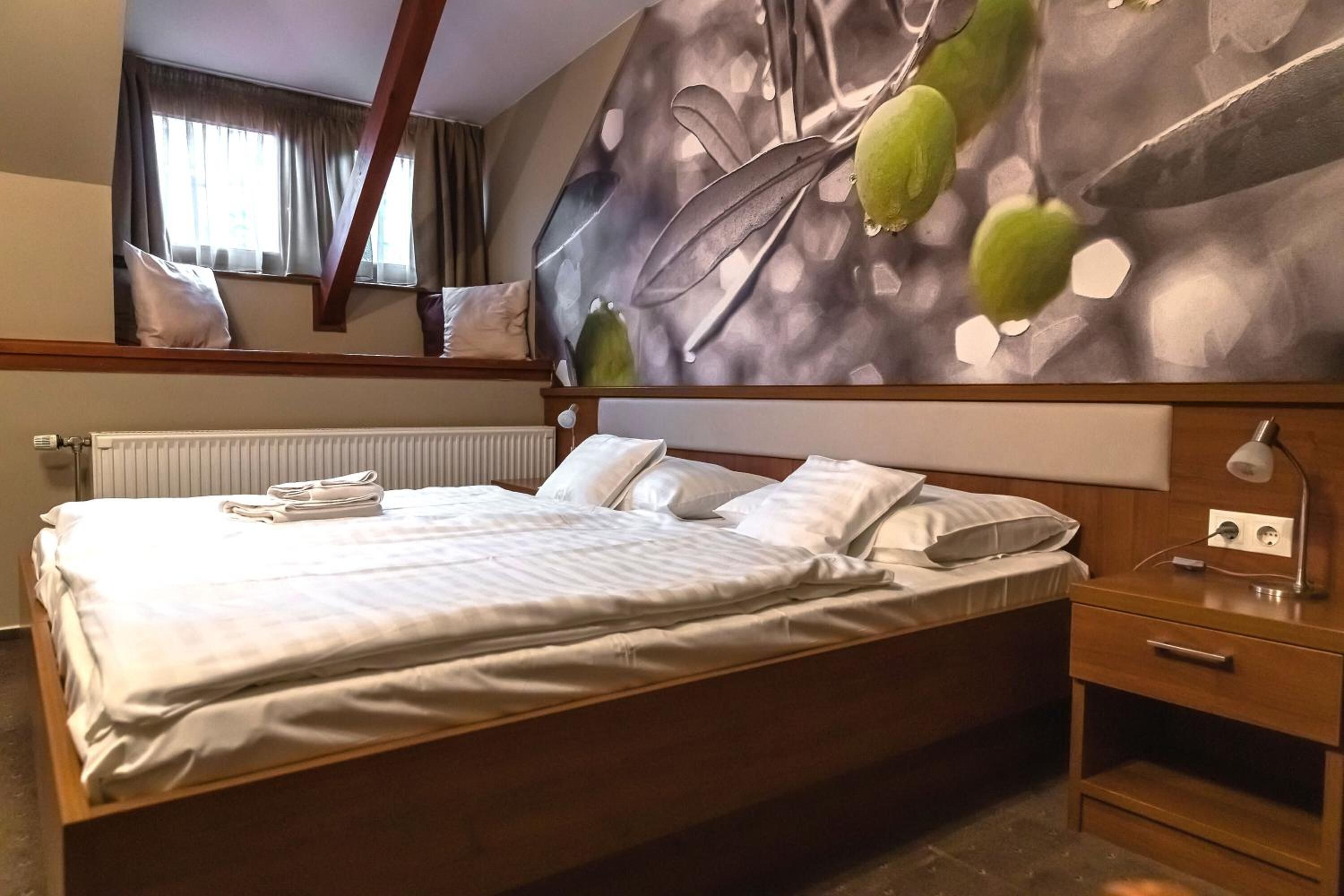 Una cama bien hecha con ropa blanca, una mesita de noche con lámpara y una pared con diseño de hojas.