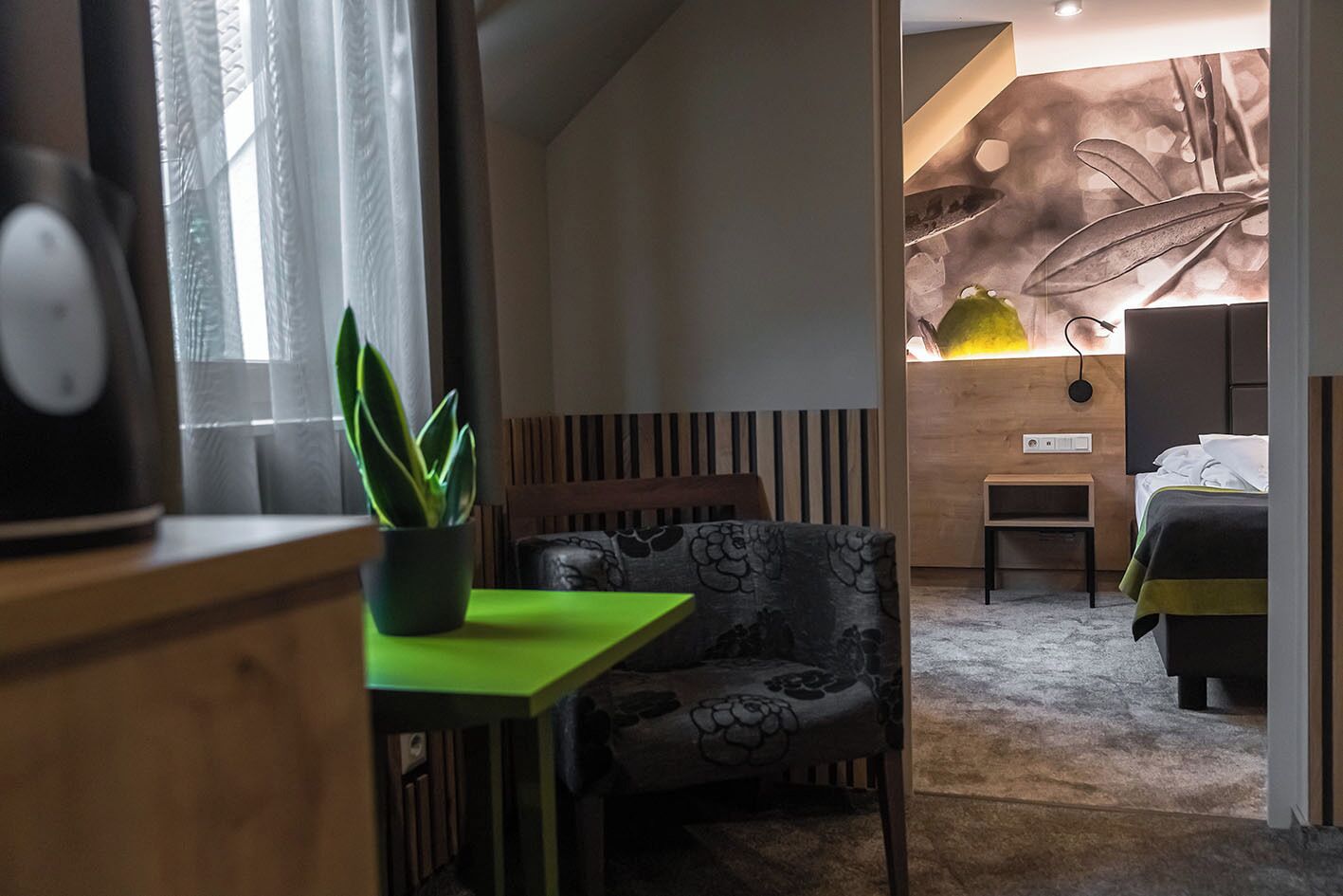 Moderni hotellihuone, jossa on puinen sängynpääty, vihreä kasvi pöydällä, kuviollinen nojatuoli ja vihreä-musta päiväpeite sängyllä.