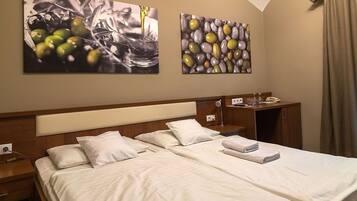 A hotel room with a double bed, two bedside tables, a nightstand, a desk with a chair, and two paintings on the wall.
