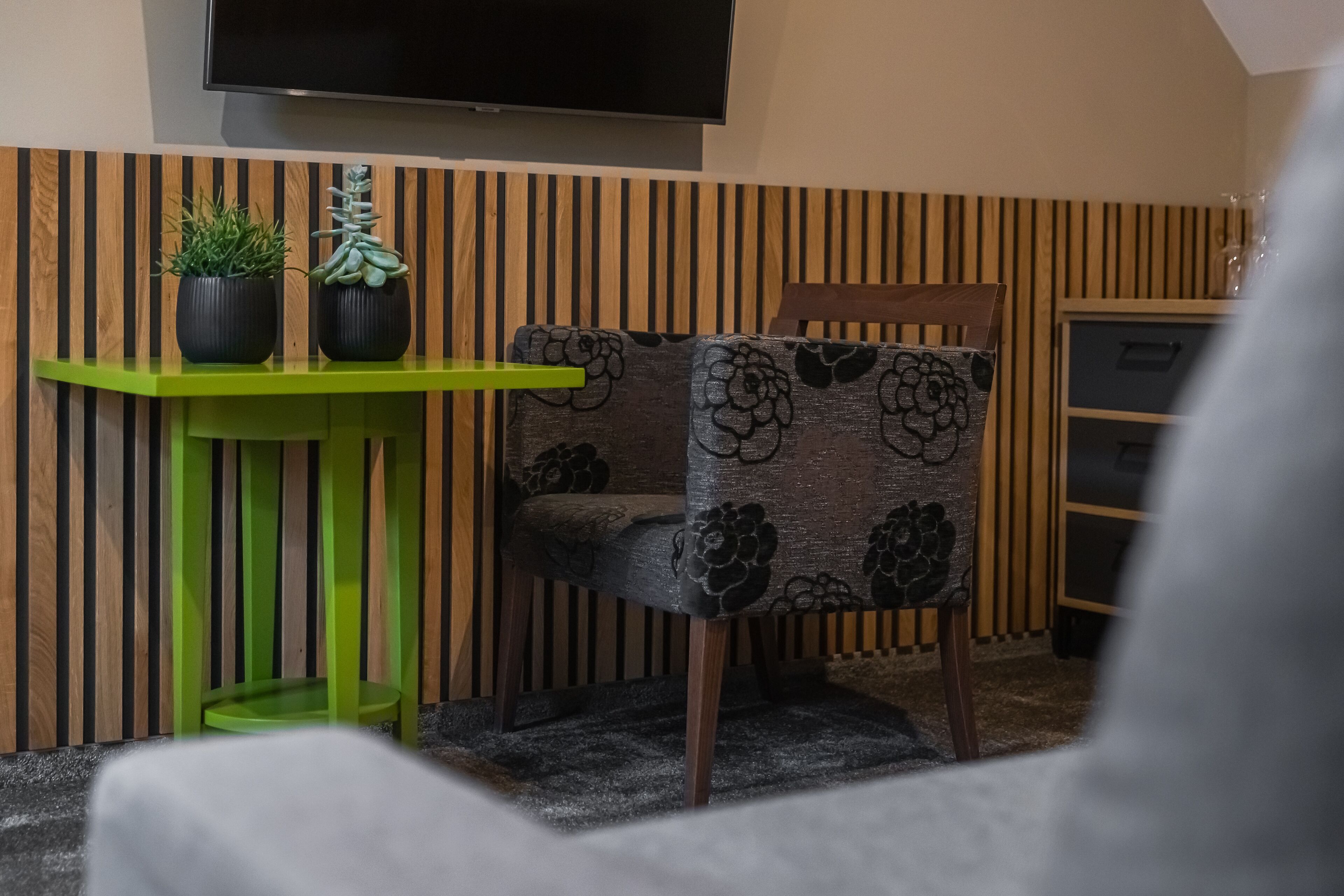 Una habitación con una pared de madera a rayas, una mesa verde con dos plantas en macetas, una silla estampada y un televisor montado arriba.