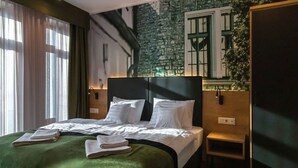 A hotel room with a large bed, a green bedspread, white pillows, a headboard, a nightstand, a wall-mounted plant, and a window with curtains.