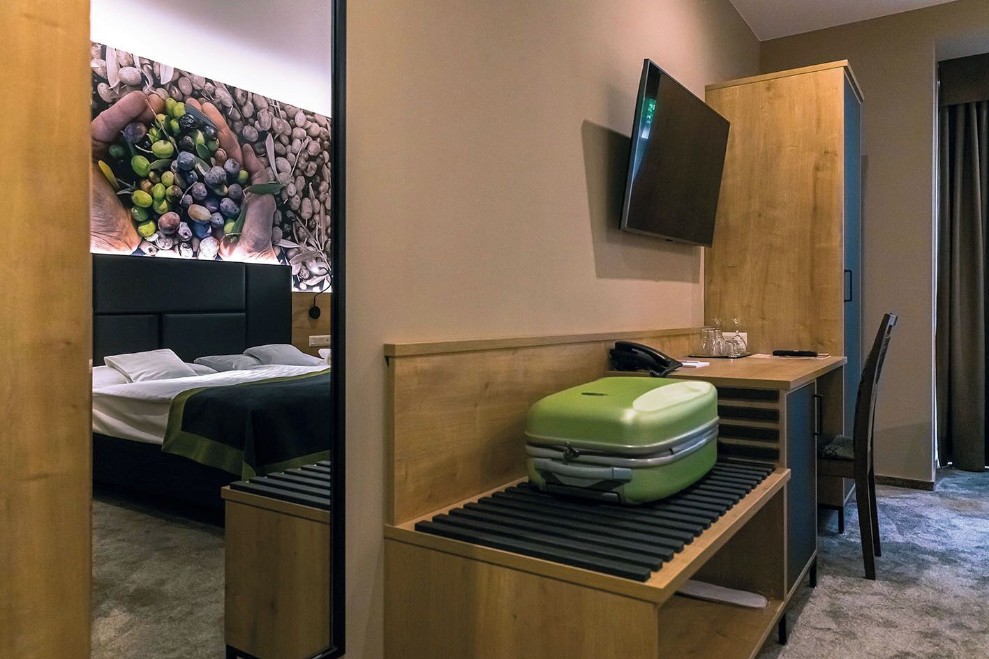Una habitación de hotel con una cama, un escritorio con una maleta, un televisor montado en la pared y una pared de piedra con un diseño de frutas.