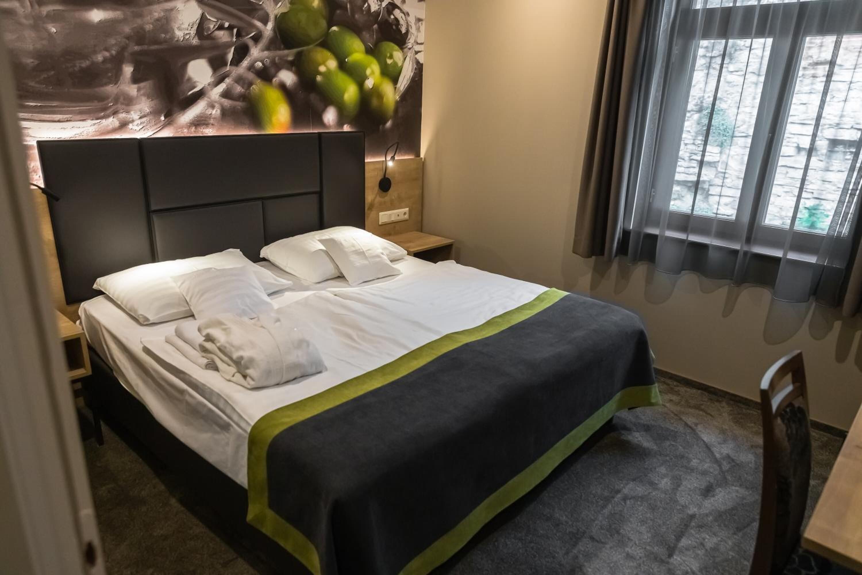 Una cama bien tendida con sábanas blancas y un detalle en verde, una mesita de noche con una lámpara y una ventana con cortinas.